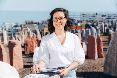 Eine Frau mit dunklen Haaren, Brille und weißer Bluse hält ein Klemmbrett in der Hand und grinst in die Kamera.