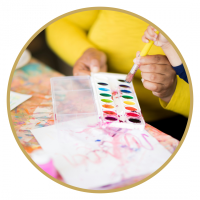 Hände beim Malen mit Pinsel und Wasserfarben an einem bunt bemalten Tisch.